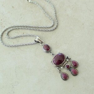 May include: A silver-toned necklace with a pendant featuring oval and teardrop-shaped, deep red gemstones. The chain is a delicate, textured design. The pendant has a cluster of stones, creating a striking visual appeal.