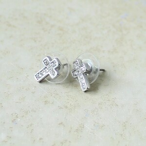 May include: A pair of silver-toned stud earrings shaped like crosses. Each earring features a polished metal frame embedded with small, clear, sparkling stones along the center. The earrings include clear, circular plastic earring backs for secure wear.