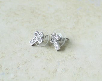 May include: A pair of silver-toned stud earrings shaped like crosses, each featuring a row of small, clear, sparkling stones embedded along the front surface and clear plastic earring backs attached to the metal posts.