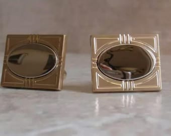 May include: A pair of square cufflinks featuring a gold-toned metal frame with geometric line detailing and a smooth, reflective, oval-shaped silver-toned center inlay. The design is symmetrical, showcasing a polished metallic finish throughout.