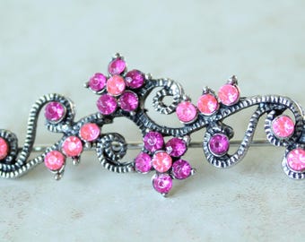 May include: A silver-toned metal brooch featuring a swirling vine design adorned with multiple round, faceted crystals in varying shades of bright pink and magenta, arranged in floral patterns and along the curved metal branches.