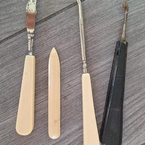 May include: A collection of antique tools, including a small knife, a pointed tool, a hook-shaped tool, and a tool with a black handle. The tools have light-colored handles and metal blades or hooks, suggesting they are for a specific craft or purpose.