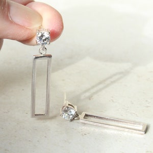 May include: Silver-toned earrings with a rectangular drop design. Each earring features a clear, round gemstone at the top, connected to a slender, open rectangle. The earrings are shown against a light background.