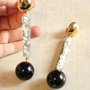 May include: A pair of drop earrings featuring a polished gold-tone dome top, a rectangular bar segment filled with shimmering silver-toned flakes, and a smooth, spherical black bead at the base, connected by small gold-toned metal jump rings.