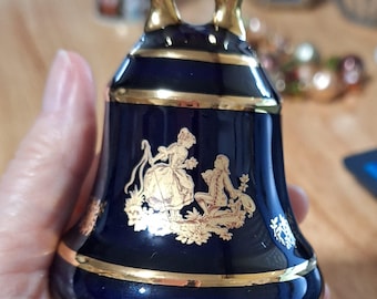 May include: A deep cobalt blue porcelain bell featuring a gold-painted scene of a person standing and another kneeling. The bell has a gold-colored handle at the top and gold trim bands around the middle and bottom edge of the bell body.