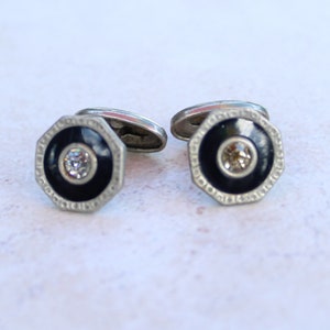 May include: A pair of silver-toned cufflinks featuring an octagonal frame with a black circular inlay and a central clear faceted stone. The metal edges have a textured pattern, and the back includes a standard swivel toggle mechanism.
