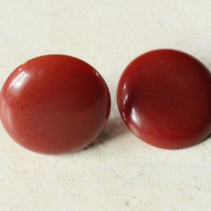 May include: Two round, dark red buttons with a glossy finish. The buttons are smooth and appear to be made of a solid material. They are set against a light, textured background.