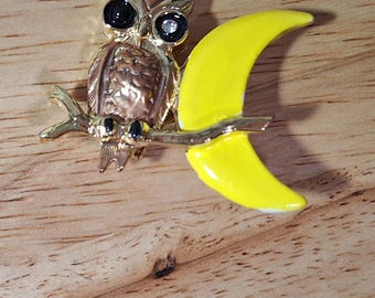 May include: A gold-toned metal brooch featuring a textured owl perched on a branch. The owl has two black circular eyes with a small clear rhinestone in the right eye. A bright yellow crescent moon shape is attached to the branch behind the owl.