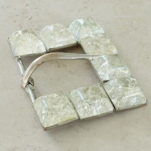 May include: A rectangular belt buckle featuring a silver-toned metal frame and seven square, translucent, crackled glass inserts. The buckle includes a curved, hinged metal prong attached to one side, designed to secure a belt strap through the frame.