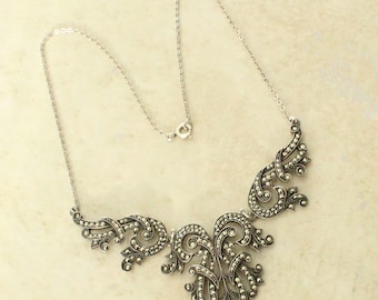 May include: A silver-toned metal necklace featuring a central ornate pendant with intricate swirling scrollwork patterns. The metal surface is densely encrusted with small, dark, reflective marcasite stones, attached to a delicate silver-toned chain.