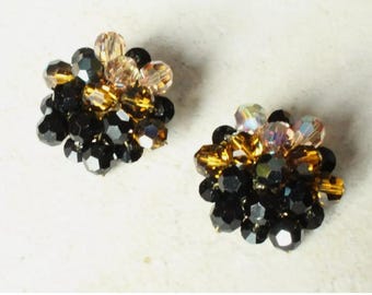 May include: A pair of cluster earrings featuring faceted beads in shades of black, amber, and iridescent clear crystal. The beads are arranged in a dense, rounded formation, creating a textured, multi-dimensional appearance for each earring.