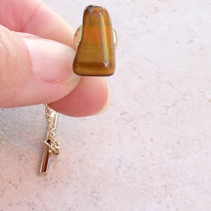 May include: A tie tack featuring a polished, irregular, golden-brown tiger's eye gemstone. The stone is mounted on a gold-tone metal base with a short, delicate gold-tone chain attached to a slender, cylindrical bar pin fastener.