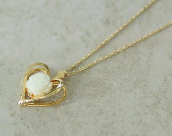 May include: A gold-toned necklace featuring a heart-shaped pendant frame. Inside the open heart frame sits a small, carved white rose. The pendant is attached to a delicate gold-toned chain with thin, interlocking oval links.