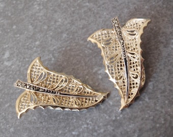 May include: A pair of gold-toned metal leaf-shaped brooches featuring an intricate openwork filigree pattern and a central spine embellished with small, dark, faceted stones that run the length of each leaf, creating a textured, metallic appearance.