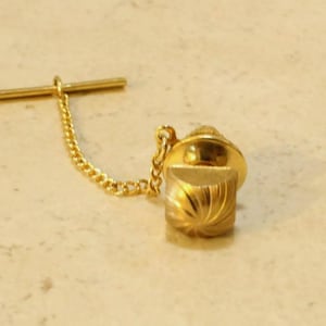 May include: A gold-colored tie tack with a decorative, spiral-carved square base and a rounded top. A delicate gold chain connects the tack to a horizontal bar. The tie tack is designed to secure a tie to a shirt.