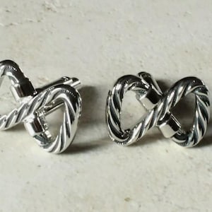 May include: A pair of silver-colored cufflinks in an infinity symbol design. The cufflinks have a twisted rope-like texture and are displayed on a light-colored, textured surface. The design is a classic accessory.