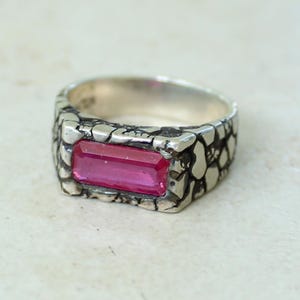 May include: A silver ring with a textured band and a rectangular, bright pink gemstone. The ring's band has a unique, irregular pattern, and the gemstone is set in a bezel setting.