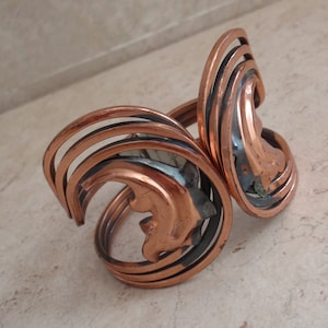 May include: A copper cuff bracelet featuring a multi-layered, spiraling wire design. The metal has a warm, metallic orange-brown finish with dark, oxidized accents between the coiled bands, creating a dimensional, open-work structure.