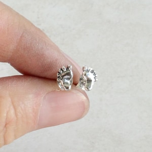 May include: A pair of small, silver-colored stud earrings. Each earring is shaped like a stylized face with detailed features. The earrings are held between fingers against a neutral background.