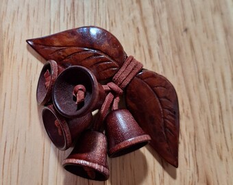 May include: A brown wooden brooch featuring two carved leaves and four small bell-shaped wooden pendants. The components are connected by thin brown leather cords, creating a cluster of natural-toned wooden elements with a polished finish.