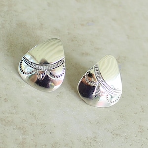 May include: A pair of silver-toned stud earrings. Each earring has a teardrop shape with a textured, ridged top and a decorative band with a detailed pattern. The earrings are displayed on a light-colored surface.
