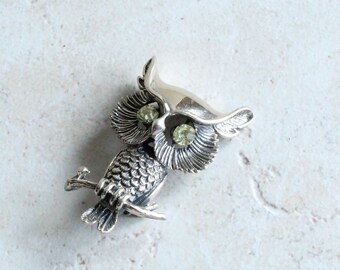 May include: A silver-toned metal owl brooch featuring detailed feather textures on the body and wings. The owl has two round, light green faceted stones set as eyes and is depicted perched on a small branch with its wings slightly raised.