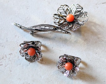 May include: A silver-toned metal jewelry set consisting of a brooch and a pair of clip-on earrings. Each piece features a filigree flower design with a central round orange bead. The brooch includes a long, slender stem with leaves.