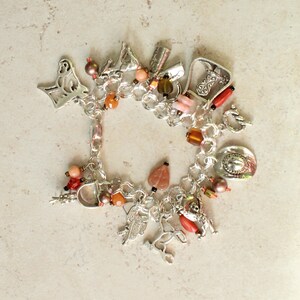 May include: A silver-tone charm bracelet featuring various western-themed metal charms including a cowboy boot, a saddle, a horse, and a cowboy hat, interspersed with round and heart-shaped beads in shades of orange, brown, and coral red.