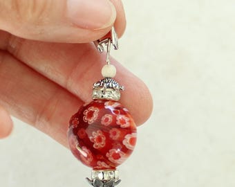 May include: A decorative charm featuring a round red glass bead with white floral patterns. The bead is accented with silver-toned metal caps, clear rhinestones, and a small white spherical bead at the top, finished with a metal lobster clasp.