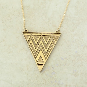 May include: A gold-toned necklace featuring a large, downward-pointing triangular pendant. The pendant has a textured surface with a repeating geometric pattern of zig-zag lines and smaller triangles, suspended from a delicate gold-toned chain.
