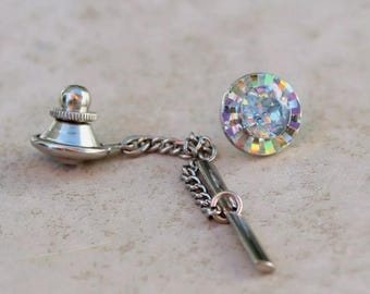 May include: A silver-toned tie tack featuring a circular head with a faceted, iridescent center stone surrounded by a reflective, multi-colored border. The piece includes a short metal chain connected to a slender bar and a secure clutch back fastener.