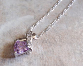 May include: A silver-toned necklace featuring a pendant with a triangular light purple gemstone. The pendant is accented by a curved silver ribbon design embedded with small clear stones, suspended from a delicate silver-toned chain.