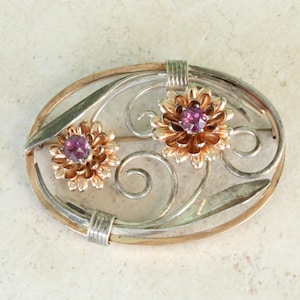 May include: Oval brooch with a silver and gold-tone design. Features two flower-shaped accents with purple gemstone centers. The brooch has a decorative, openwork design with swirling lines and leaf accents.