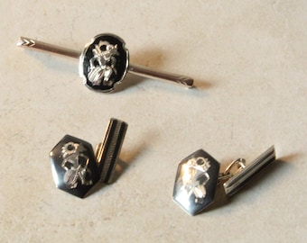 May include: A silver-toned tie bar and a pair of matching cufflinks, each featuring a black inlay with a detailed engraved figure. The tie bar has an oval center, while the cufflinks have a hexagonal shape with a hinged bar attachment.