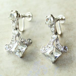 May include: A pair of silver-toned clip-on earrings. Each earring features a large, square, clear crystal at the bottom, with smaller crystals and decorative flourishes. The earrings have a vintage, Art Deco style.