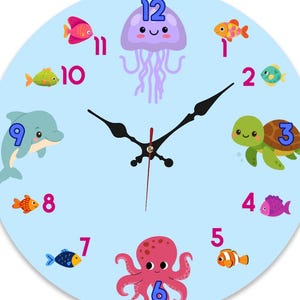 May include: A round wall clock with a light blue face featuring colorful cartoon sea creatures including a purple jellyfish, a green turtle, a blue dolphin, and an octopus. The numbers 1 through 12 are printed in pink and blue around the dial.