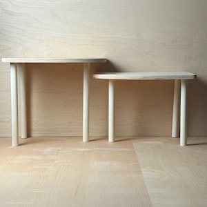 May include: Two small, light-colored wooden tables. Each table has a rectangular top and three cylindrical legs. The tables are different sizes, with one slightly larger than the other. The background is a light wood panel.