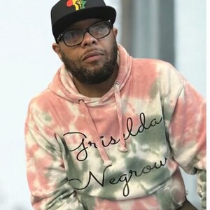 Op de afbeelding: Een tie-dye hoodie in roze, groen en wit, met de tekst "Griselda Negrow" in zwart schrift. De persoon draagt een zwarte baseballcap met een kleurrijk vuistlogo en een bril met zwart montuur.