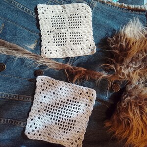 Peut inclure: Deux patchs carrés en crochet blanc avec des motifs ajourés reposent sur une veste en jean. La veste a des boutons en métal et des coutures visibles. Une garniture en fourrure marron et une longue plume fine marron sont également présentes.