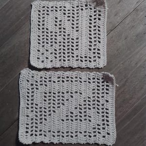 Peut inclure: Deux pièces carrées en crochet blanc cassé avec un motif ajouré répétitif. Le travail au crochet a un aspect texturé, et les pièces sont posées sur une surface en bois. Le design ouvert suggère un textile décoratif ou fonctionnel.