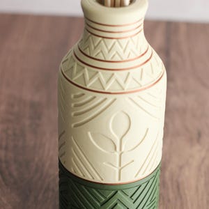 May include: A decorative ceramic vase with a cream-colored upper section and a green base. The cream section features etched geometric and floral patterns with copper-colored accents. The vase holds several light-colored reeds.