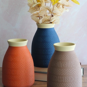 Puede incluir: Tres jarrones de cerámica en tonos naranja, azul y marrón, cada uno con un borde crema y un patrón geométrico. El jarrón azul contiene un ramo de flores secas amarillas y blancas. Un accesorio decorativo para el hogar.