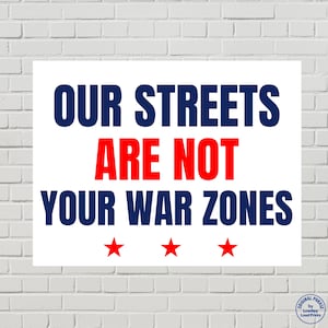 May include: White rectangular sign with the text "OUR STREETS ARE NOT YOUR WAR ZONES" in navy blue and red. Three red stars are at the bottom. The sign is against a white brick wall.