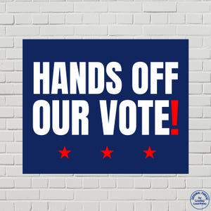 May include: A navy blue rectangular sign with white text that reads "HANDS OFF OUR VOTE!" and three red stars at the bottom. The exclamation point is red. The sign is against a white brick wall.