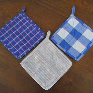 May include: Three quilted pot holders in a variety of patterns. One is blue and pink plaid, another is blue and white checkered, and the third is white with blue stripes. Each has a loop for hanging.