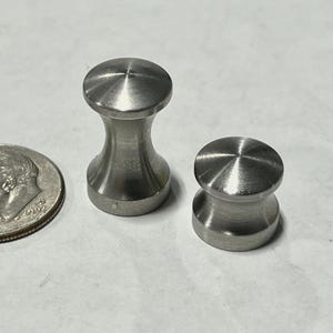 May include: Two small, brushed metal knobs with a rounded top and bottom, connected by a narrower, curved midsection. The knobs are next to a U.S. dime for size comparison. The knobs are likely for furniture or cabinetry.