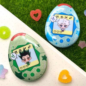 Puede incluir: Dos juguetes digitales para mascotas con forma de huevo y brillantes. Uno es de color verde azulado con una ilustración de personaje, el otro es verde con un personaje diferente. Ambos tienen una parte superior roja y una pantalla amarilla. Los juguetes están sobre una superficie blanca con hierba verde y caramelos.