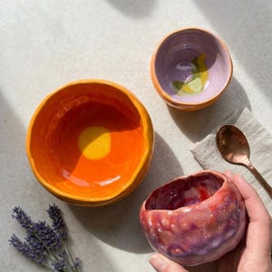 Puede incluir: Tres cuencos de cerámica hechos a mano en colores vibrantes. El cuenco más grande es naranja con un centro amarillo. Un cuenco más pequeño es morado con un diseño de limón, y otro es rojo con motas blancas. También hay una cuchara de cobre y una ramita de lavanda.