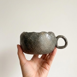 May include: A handmade, speckled ceramic mug with a dark gray exterior and a rounded handle. The rim of the mug has a wavy, organic shape. The mug is held in a hand against a plain white background. The mug is designed for beverages.