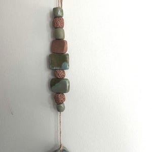 Puede incluir: Móvil colgante de cerámica con una cadena de cuentas verdes, marrones y rosas. Las cuentas están conectadas por una cuerda natural y terminan con un anillo retorcido azul y gris con un acento de hoja dorada.